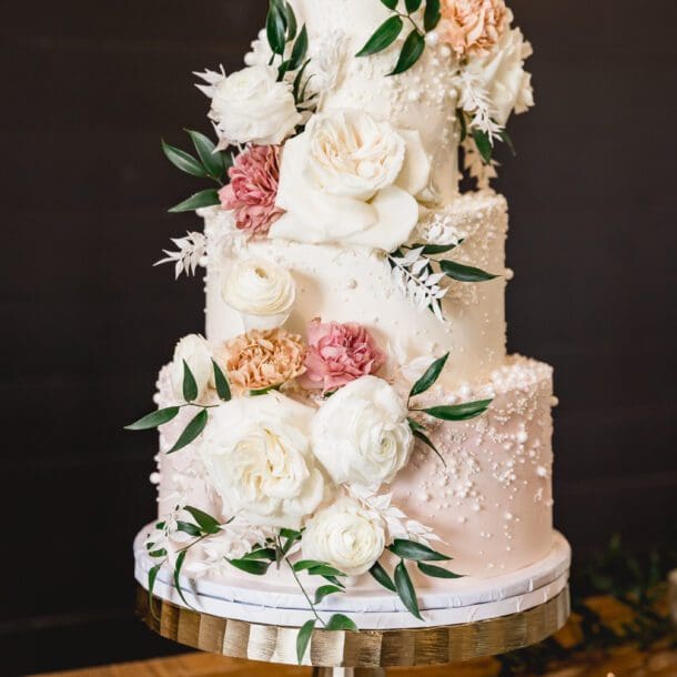 Elegant three-tier wedding cake with white and blush floral decorations on a gold stand photo by Bushel + Peck Photo