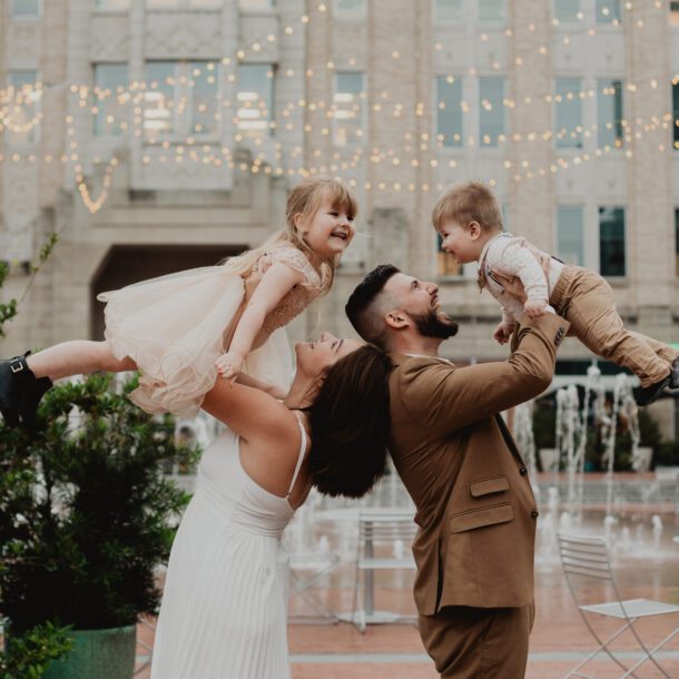 Happy family portrait session with parents lifting children under string lights in city square — Bushel + Peck Photo.