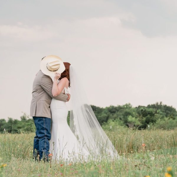 Romantic Wedding Photo in East Texas Photography Location Bride and groom share a romantic kiss in an East Texas field, with the groom’s cowboy hat playfully shielding their faces. The bride’s flowing veil and the rustic landscape highlight this charming East Texas photography location.