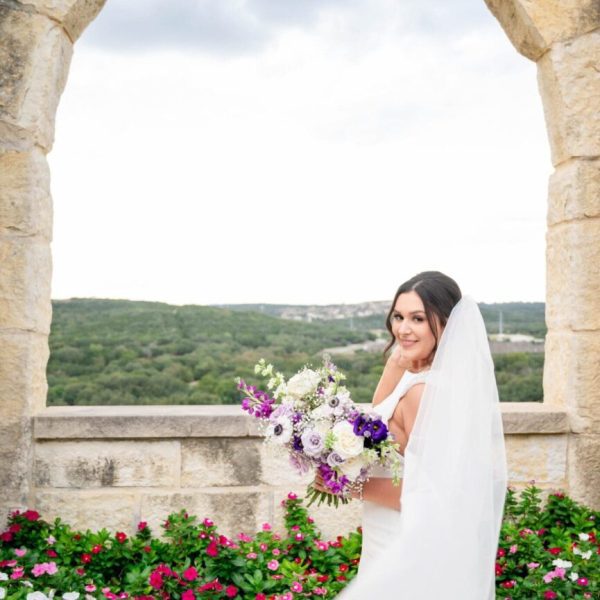 Bridal beauty in South Central Texas — a stunning portrait at a picturesque photography location near San Antonio, Austin, and Houston. Smiling bride posing with purple and white bouquet at a scenic South Central Texas photography location near San Antonio, Austin, and Houston, framed by stone arches and rolling hills.